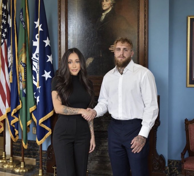 Jake Paul and his partner Brittany Renner pose together in what appears to be a formal government setting, with Paul shaking her hand in front of a historical portrait and American flags, suggesting a meeting with an official or diplomatic engagement. The image relates to the post about a challenging month by hinting at a significant or perhaps unexpected encounter amid personal or professional turbulence.