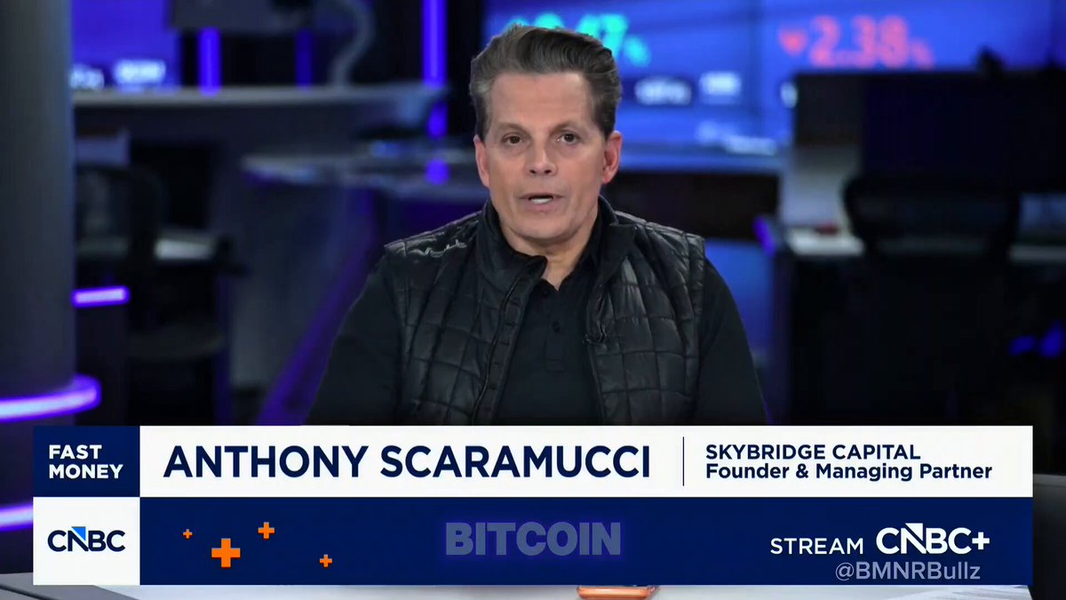 The image features a man wearing a black vest, sitting at a desk with a computer monitor behind him. The man appears to be talking about Bitcoin on a news program or a TV show. He is dressed in a black shirt and has short hair. The background of the image shows a blue wall with a clock on it, indicating that the scene takes place during daytime hours.