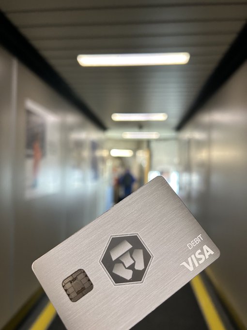 A metallic Crypto.com Visa debit card is held up in the foreground, with the words "DEBIT" and "VISA" visible, set against the blurred backdrop of an airport jet bridge. The image ties into the "#cryptoirl #crofam" caption by showcasing the real-world utility of using a crypto-branded Visa card for everyday travel expenses.
