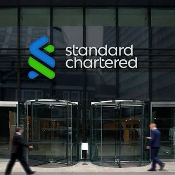 The image shows the entrance of a Standard Chartered Bank building, featuring the bank's distinctive blue and green logo alongside its name displayed on the glass facade, with two businessmen walking past the revolving doors. This visual reinforces the post's bullish Bitcoin outlook, as Standard Chartered—the financial institution featured—has notably raised its price target, suggesting $100,000 may be conservative given strengthening institutional demand.