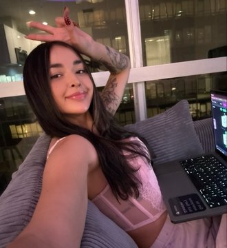 A woman with long dark hair posing with her arm raised near her head, sitting on a couch next to an open laptop.