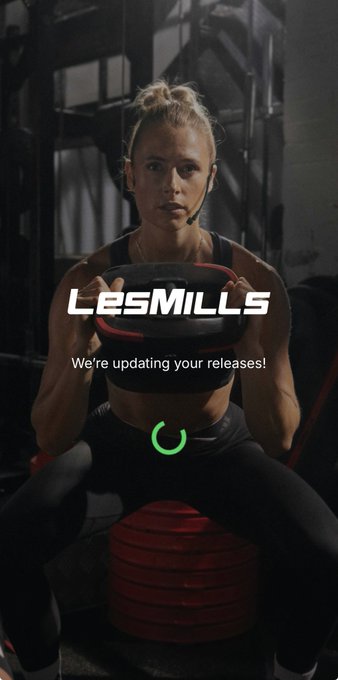 A woman with a headset performs a squat holding a weight plate, overlaid with the text "LES MILLS" and "We're updating your releases!" with a loading icon.