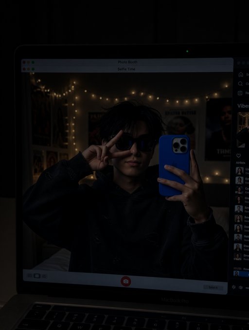 A moody mirror selfie taken on a MacBook Pro Photo Booth, showing a person in a black hoodie and sunglasses posing with a peace sign while holding a blue iPhone, with string lights and posters in the background.