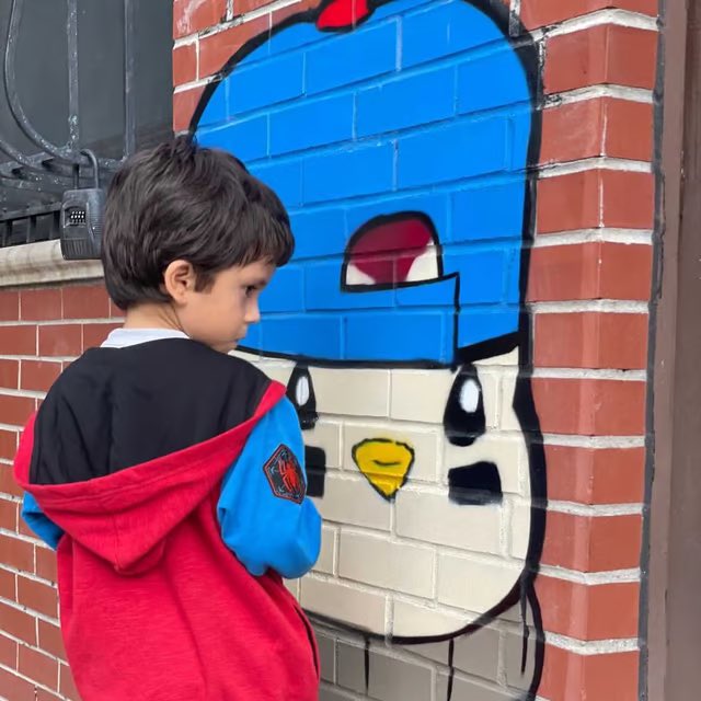 A young boy in a red and blue hoodie stands admiringly next to a colorful Pudgy Penguin mural painted on a brick wall, reflecting the post's sentiment about the next generation connecting with the iconic Web3 penguin brand.