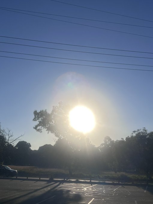 A scenic photograph of a bright sun setting behind silhouetted trees, with power lines crossing the blue sky and an empty parking lot visible in the foreground.