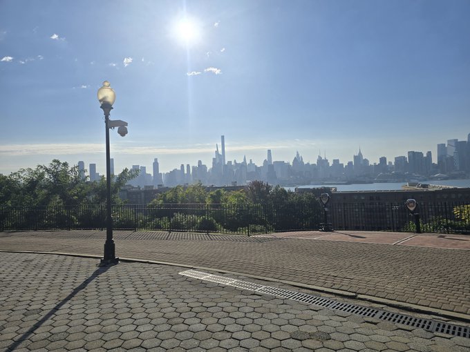 A sunlit promenade overlooks the New York City skyline across the water, with a classic lamppost standing prominently in the foreground. The serene, hopeful atmosphere of the cityscape complements the uplifting Saturday message of blessings and love.