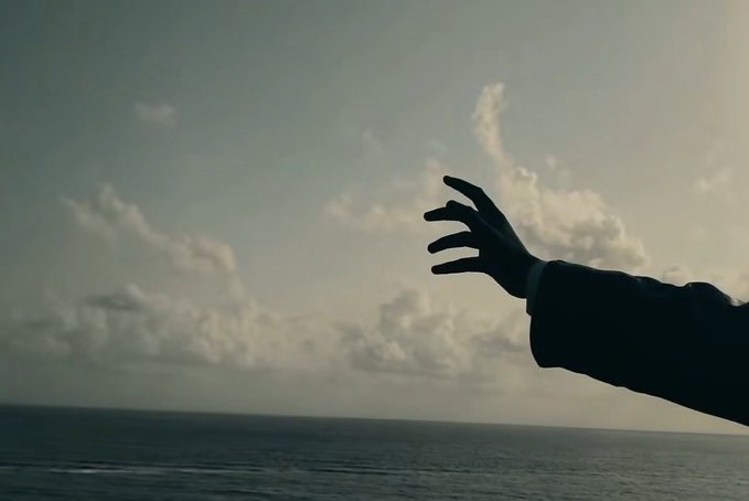 A silhouette of a hand reaching out toward a cloudy sky over the ocean, evoking a sense of yearning or longing.