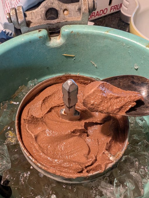 A close-up photo of a traditional Mexican stone metate grinding chocolate, showing a thick brown chocolate paste being worked around the central stone pestle, surrounded by ice in a green ceramic bowl.