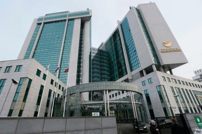 The image shows the modern glass-and-steel headquarters building of Sberbank, with its distinctive logo visible on the upper right corner and a Russian flag flying near the entrance. This relates to the post as it depicts the physical corporate headquarters of Sberbank, Russia's largest state-owned bank, which is reportedly preparing to enter the cryptocurrency market with Bitcoin trading and custody services.