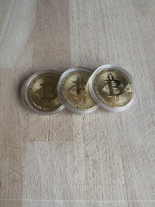 Three gold-colored Bitcoin commemorative coins in clear plastic casings are fanned out on a light wooden surface.