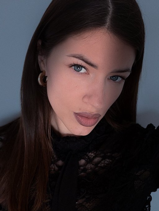 The image features a woman with long brown hair wearing a black top. She has her eyes closed and is looking directly at the camera. The background of the image is white, which contrasts with the dark tones of the woman's hair and outfit. There are no other objects or texts in the image, making it a simple yet striking composition that highlights the subject.