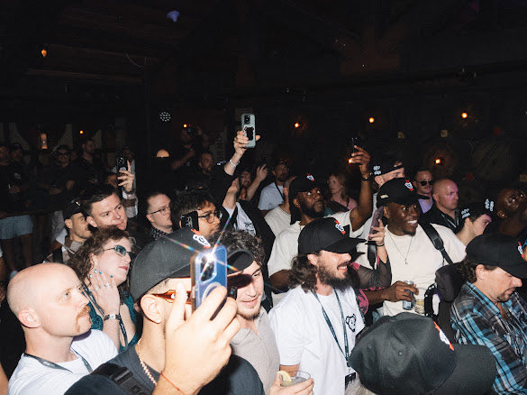 A crowded concert or event scene with many attendees holding up cameras and phones, some wearing caps and casual clothing, in a dimly lit venue.