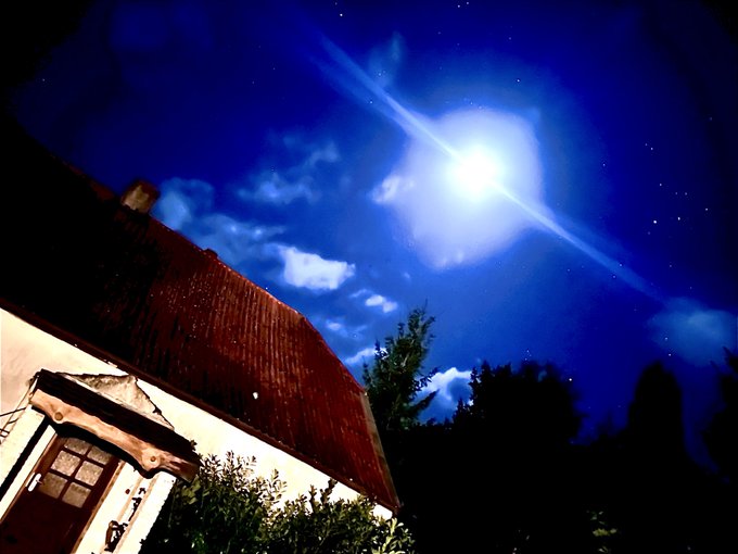 A nighttime photograph looking up at a brilliantly shining moon in a dark blue sky with scattered clouds, positioned above the dark silhouette of a house roof and trees.