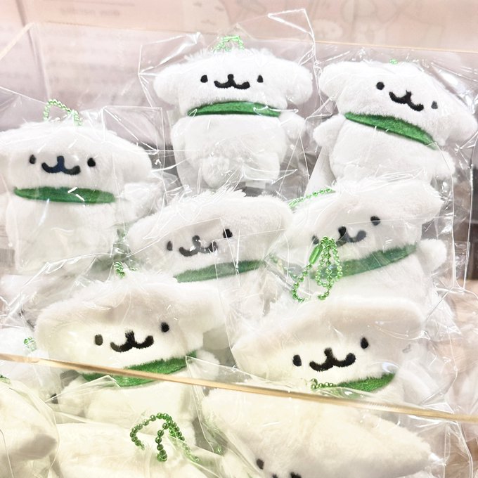 A collection of cute white plush toys with simple embroidered faces and green collars, each packaged in clear plastic bags with green ball chains, displayed in an acrylic container for the #NERDOG popup event.