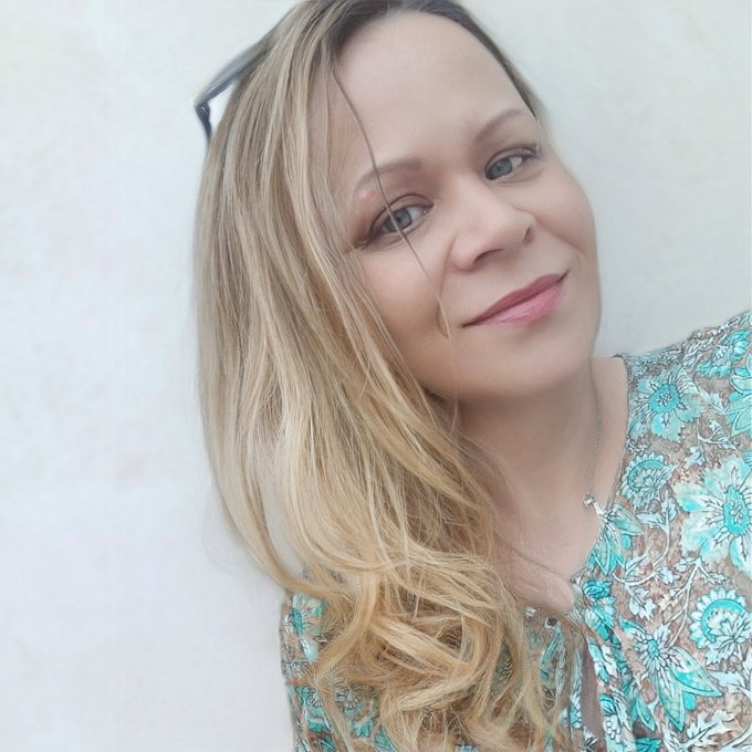 The image features a woman with blonde hair wearing a blue floral shirt, smiling at the camera while looking directly at it. The background of the photo is white, which contrasts nicely with her outfit and highlights her presence. The caption above the image reads "GM fam BTC is holding its nerve. SOL is trying to remind everyone why people love it. NFTs still refuse to die, even though the internet has buried them more times than some celebrities." This text seems unrelated to the woman in the photo but might be a reference or an inside joke between her and someone else who shared this image on social media.