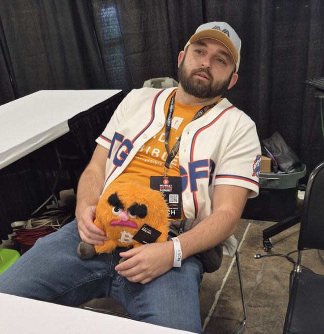 A bearded man wearing a baseball cap and baseball jersey sits at a table holding an orange plush toy at what appears to be a gaming convention.