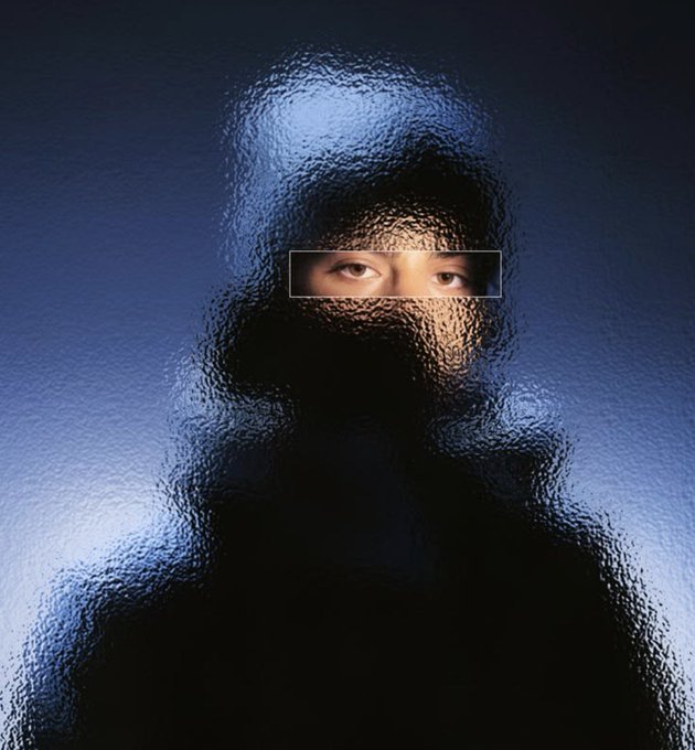 A person's eyes are visible through a narrow horizontal slit in a dark, textured surface that covers the rest of their face, set against a blue background.