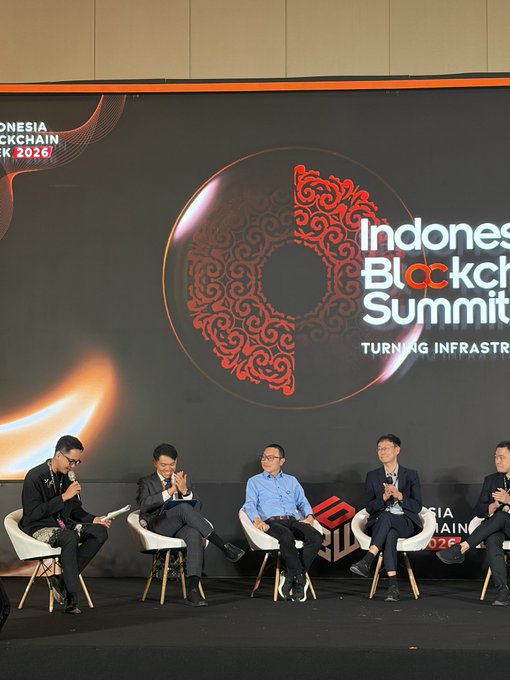 A panel of five men sits on stage at the "Indonesia Blockchain Summit" in 2026, with one speaking into a microphone while the large screen behind displays the event's branding and tagline "Turning Infrastructure." The summit setting aligns with the post's discussion on crypto's new legal status, featuring speakers addressing the transformation of cryptocurrency from commodity to recognized capital in Indonesia.