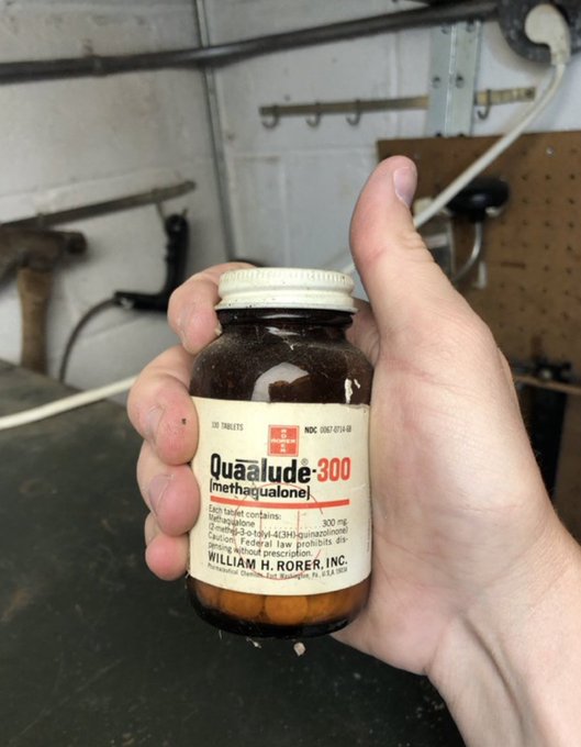 A hand holds a vintage brown glass bottle labeled 'Quaālude-300 (methaqualone)' by William H. Rorer, Inc., containing 300 mg tablets, against a workshop wall with tools.