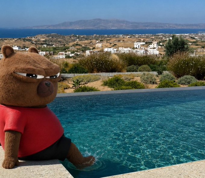 A plush brown bear toy wearing a red shirt and black shorts sits at the edge of a swimming pool, dipping its feet into the water with a scenic coastal landscape and blue sea in the background.