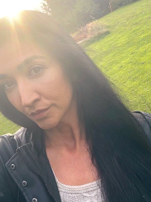 A woman with long dark hair takes a golden-hour selfie in a grassy park, wearing a black leather jacket over a white top, capturing a serene morning vibe that fits the "GM" (Good Morning) $XRP greeting and the motivational message about locking in for the day.