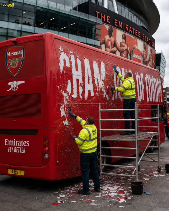The image shows two men working together to paint a red bus, possibly an Emirates Fly Better bus or a bus from the Emirates Stadium. The men are wearing yellow jackets while they work on the bus. They seem to be engaged in their task, as there is a bucket of white paint nearby that has been used for painting the bus.