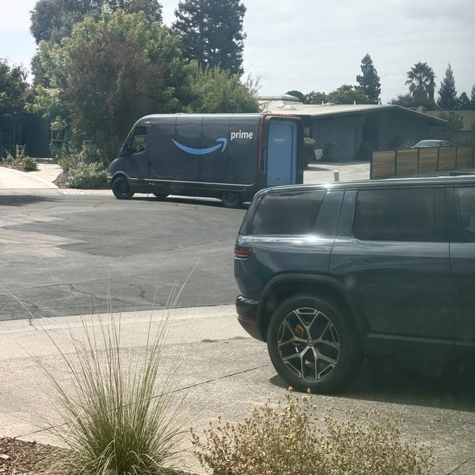 An Amazon Prime delivery van is parked on a residential street in front of a house, with the recognizable 'prime' logo and blue smile arrow visible on its side, while a dark-colored SUV with sleek alloy wheels is parked in the foreground driveway near some ornamental grasses.