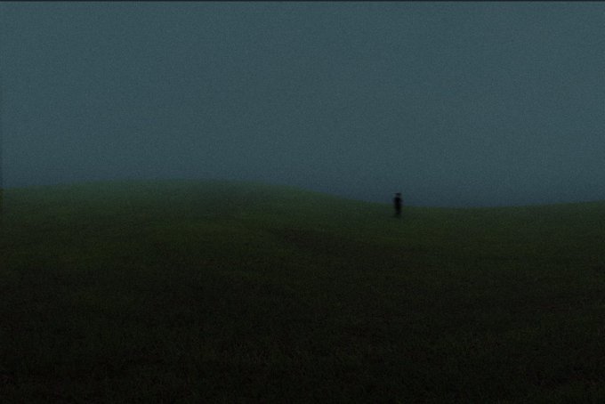 A dark, moody photograph showing a lone silhouetted figure standing in a grassy field under a dim, overcast sky.