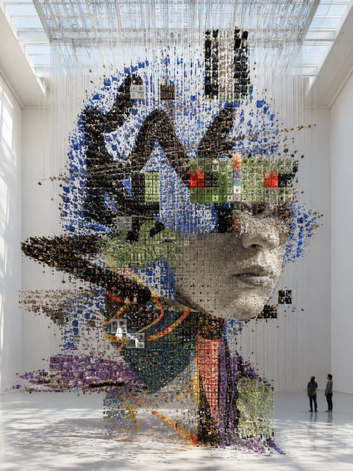 A large-scale art installation in a bright white gallery space features a massive floating portrait of a woman's face constructed from thousands of suspended colorful cubes and pixel-like elements, with two visitors standing nearby for scale.