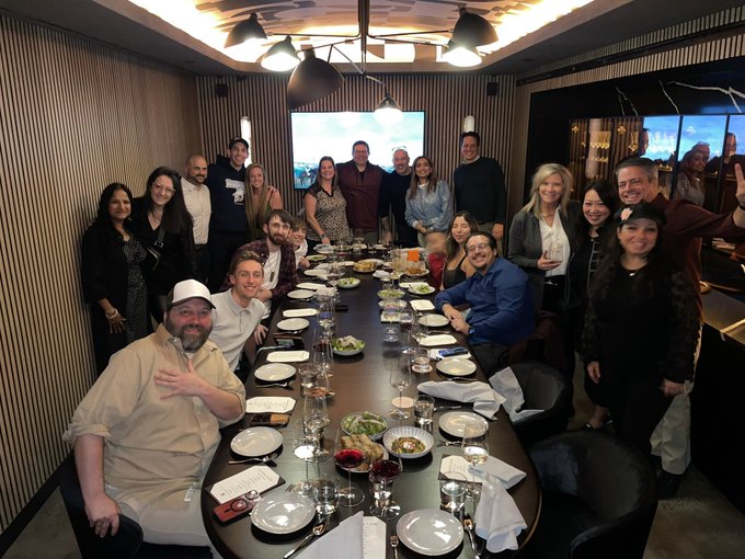 A large group of people smiling and posing for a photo around a long dining table in a dimly lit private dining room.