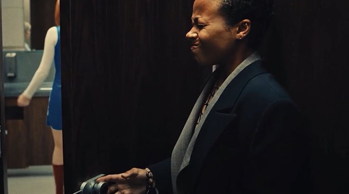 A woman in a dark blazer stands in a dimly lit room, smiling while holding an object, with another person in a blue dress visible in the background.