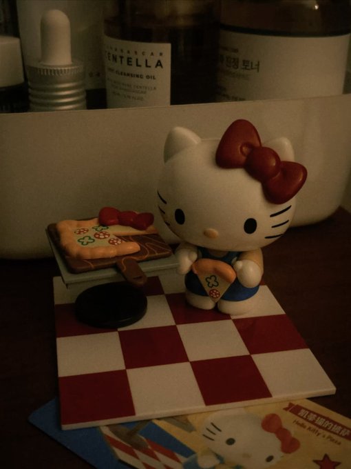 A small Hello Kitty figurine sits on a red and white checkered surface, holding a paintbrush next to a miniature palette with red paint. Bottles labeled 'CENTELLA' and other skincare products are blurred in the background.