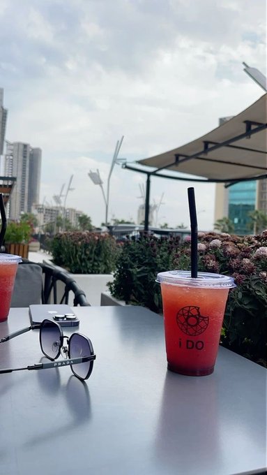 A real photograph showing a plastic cup with a red drink, a black straw, sunglasses, and a smartphone resting on a white table at an outdoor café, with the text 'i DO' visible on the cup.