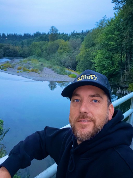 A selfie of a man with a beard wearing a black hoodie and a dark baseball cap with gold lettering, standing on a bridge overlooking a river surrounded by dense green trees at dusk.