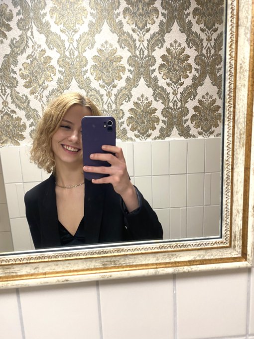 A blonde woman wearing a black blazer takes a mirror selfie with a purple smartphone, smiling brightly in front of a wall with damask-patterned wallpaper and white tiles.