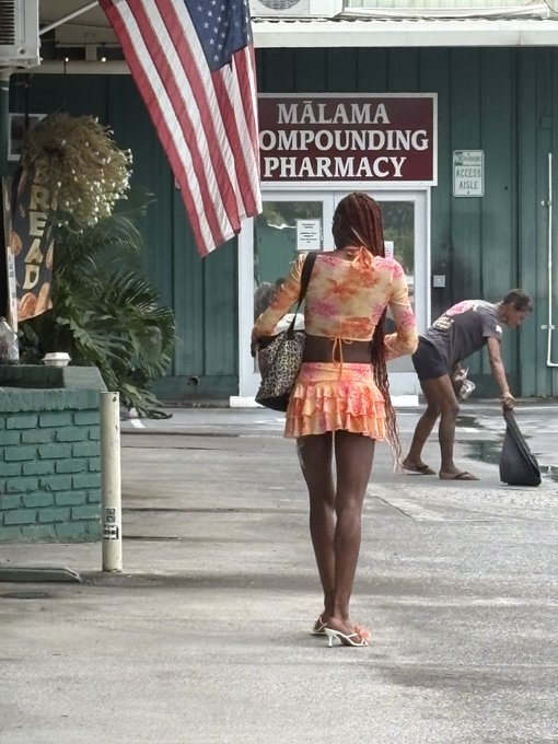 A woman in an orange floral top and skirt walking past a 'MĀLAMA COMPOUNDING PHARMACY' sign with an American flag, while another person bends over in the background.