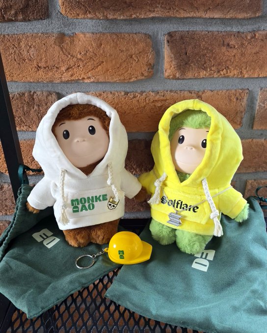 Two plush mascots in hoodies sit on green pouches against a brick wall — a brown monkey-like figure in a white 'MONKE DAO' hoodie and a green Solflare character in a yellow hoodie, with a small yellow hard hat keychain in front.