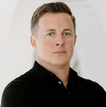 A man with short brown hair wearing a black polo shirt looks thoughtfully toward the camera against a blurred white background. The image appears to be a portrait of Elon Musk, suggesting a connection to his technology ventures and the ecosystem of transactions referenced in the post.