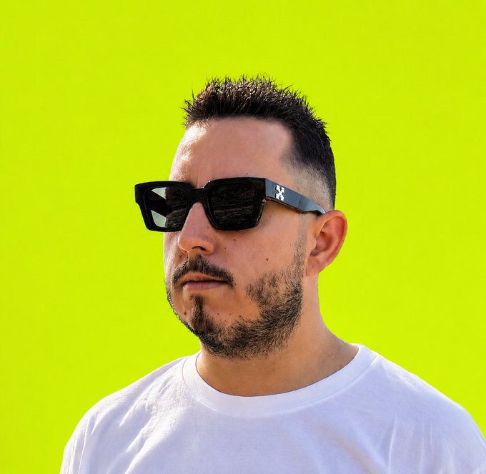 Portrait of a bearded man wearing black sunglasses and a white t-shirt against a bright yellow-green background.