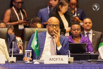 The image shows a man sitting at a table with a green, white, and black flag behind him. The man appears to be wearing glasses and has his hand over his mouth as if deep in thought or contemplation. In the background, there are other people present who may also be engaged in discussions or meetings.