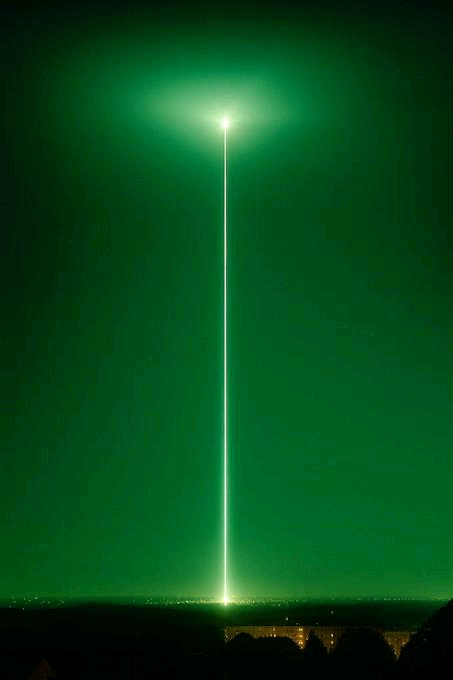 A bright green laser beam shoots vertically into the night sky from a hilltop vantage point above a city, creating a luminous beam with a halo at its tip.