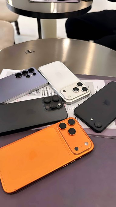 Five iPhones in various colors (blue, white, black, and orange) with distinctive camera setups are arranged on a metallic table, likely as part of a giveaway challenge to guess which phone belongs to which participant in exchange for 1 SOL.