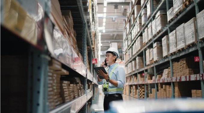 A warehouse worker wearing a hard hat and safety vest stands between tall shelves, holding a tablet and speaking into an earpiece.