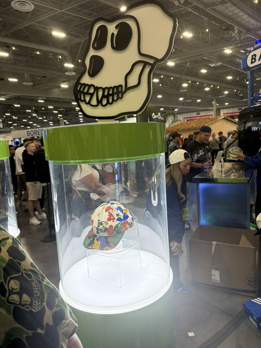 A convention display featuring a large cartoon skull logo above a clear cylindrical case containing a colorful patterned baseball cap.