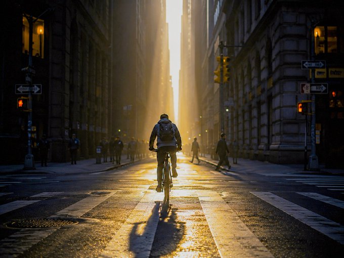 A lone cyclist rides toward a glowing sunrise between towering urban buildings, casting a long shadow across the crosswalk, while nearby street signs read "ONE WAY." The dramatic golden light perfectly captures the "Morning Light" theme of the artwork, posted as a 1/1 edition NFT on Tezos/Objkt.