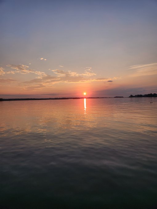 A serene sunset over a calm body of water, with the sun sitting just above the horizon, casting a warm orange and golden glow across the sky and reflecting on the water's surface.