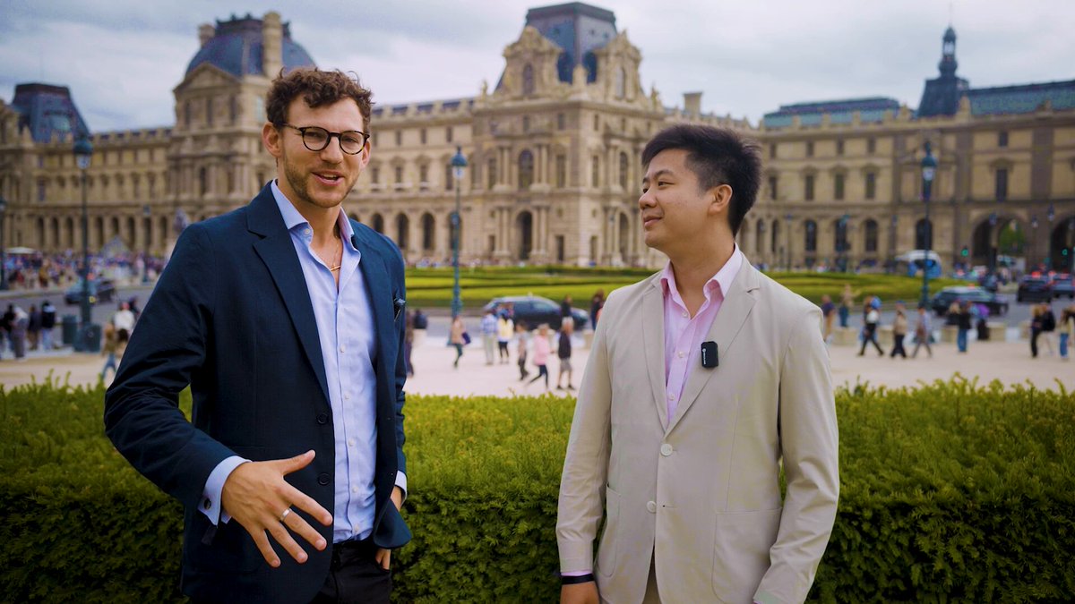 The image shows two men standing next to each other near a building with a large courtyard. One of them has a camera around his neck, suggesting they might be tourists exploring the city together. They are both dressed professionally, wearing suits and ties. The backdrop features a beautiful view of the Palace of Versailles in Paris, France, which adds an elegant atmosphere to their conversation.