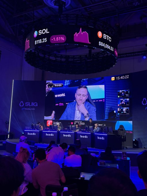 A large digital display board hangs above a conference stage showing crypto prices including SOL at $118.25 (-1.51%) and BTC at $84,084.53, while a giant screen below features speakers at the "SUI Basecamp" event, with "hud1" branding visible on multiple monitors and $1,000 prize amounts displayed, capturing the competitive trading atmosphere mentioned in the post.