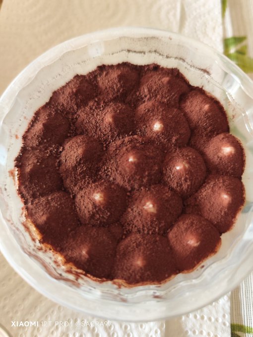 The image shows a close-up view of a dessert, possibly a Tiramisu or similar dish, served in a glass bowl with a white lid. The dessert appears to be made from chocolate and has a rich brown color, suggesting its delicious taste. The image also contains text that reads "Tiramisú que delicia #hive". This could potentially be a reference to the post on social media or an advertisement for the dessert.