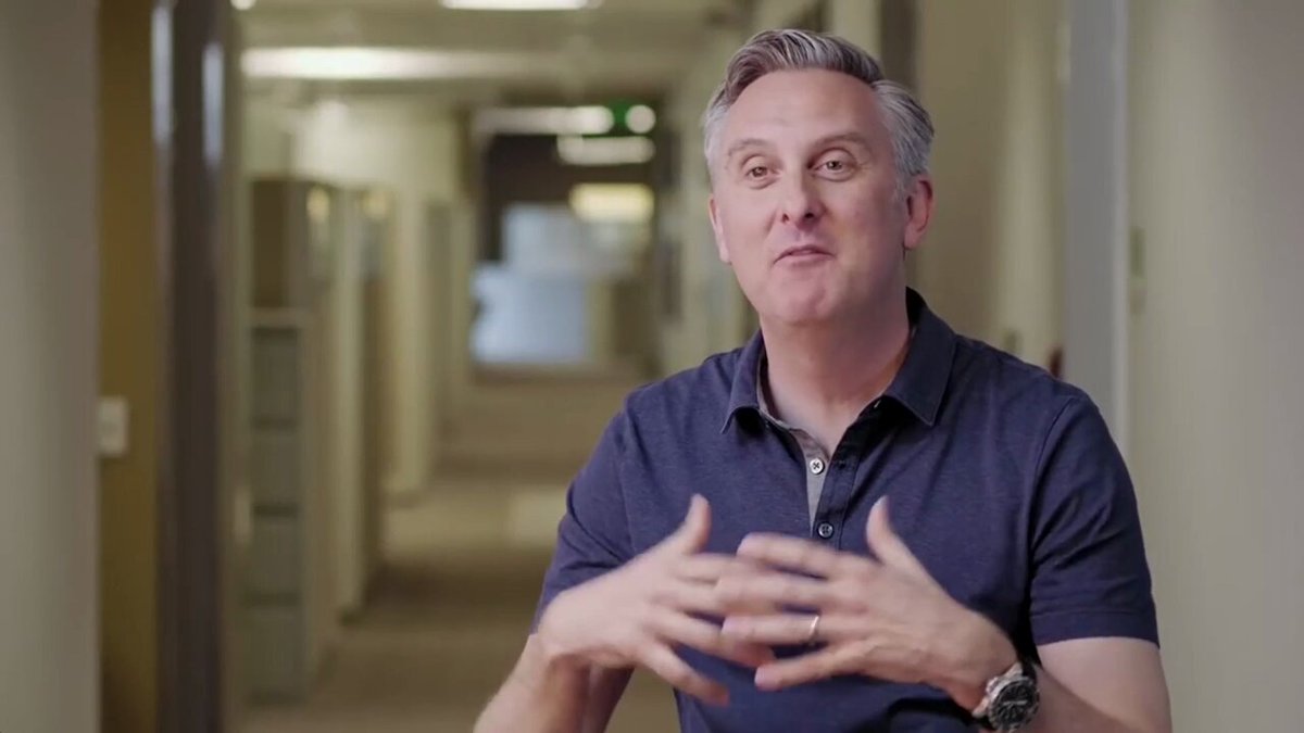 The image features a man wearing a blue shirt, sitting down and talking into a microphone. The background of the image shows an office hallway filled with cubicles and desks, suggesting that he might be at work or participating in a meeting. There is also text on the image that reads "watch IBM, one of the world's largest tech leaders state why they chose $XLM IBM are trending right now as their stock is flying! They have partnered with @StellarOrg many times over the years!"
